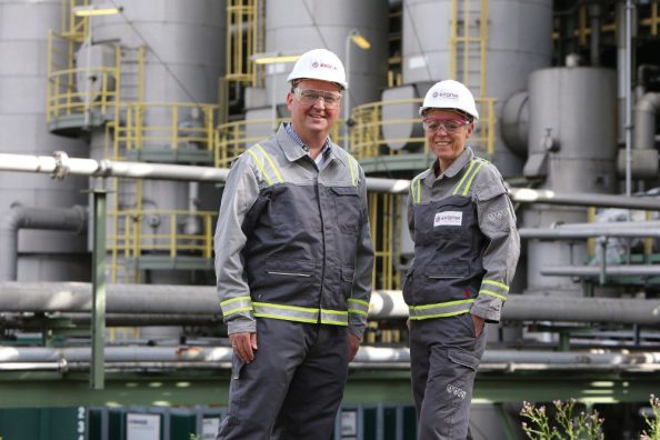 Ein Mann und eine Frau in Schutzkleidung stehen vor einer Fabrikanlage.