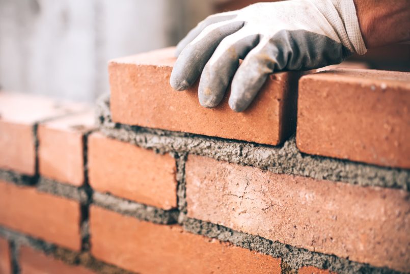 Hand mit Schutzhandschuh legt einen Zieglstein auf eine Mauer mit frischem Mörtel.